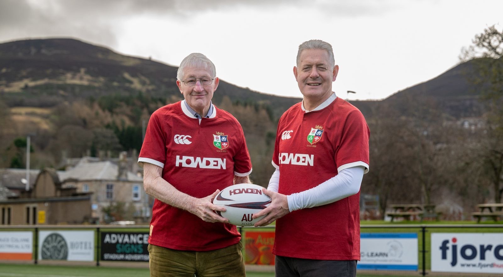 Lions legends Jim Telfer and Gavin Hastings, in Howden-branded jerseys and a rugby ball, in front of the Eildon Hills